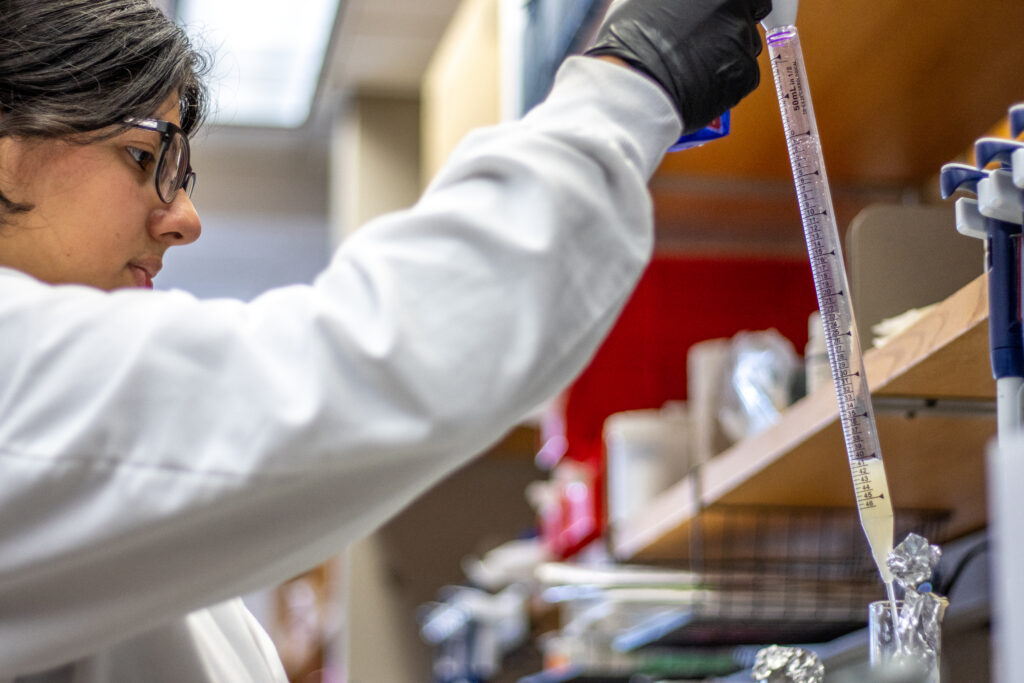 Woman in lab coat pours liquid out of pipette into flask.