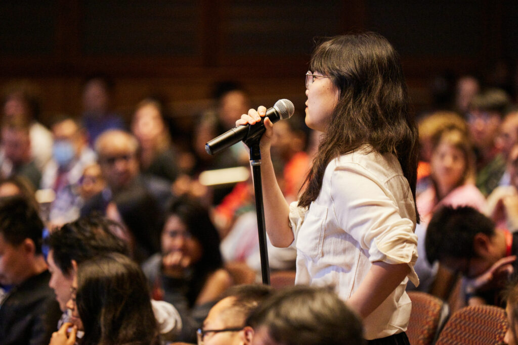 Woman asks a question into a microphone in a full auditorium at IPI Surfacing.