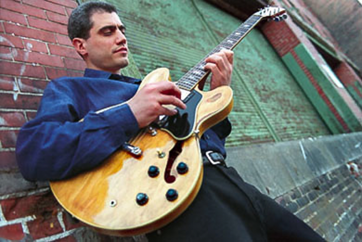 Issi Rozen playing his guitar and leaning against a distressed brick wall.