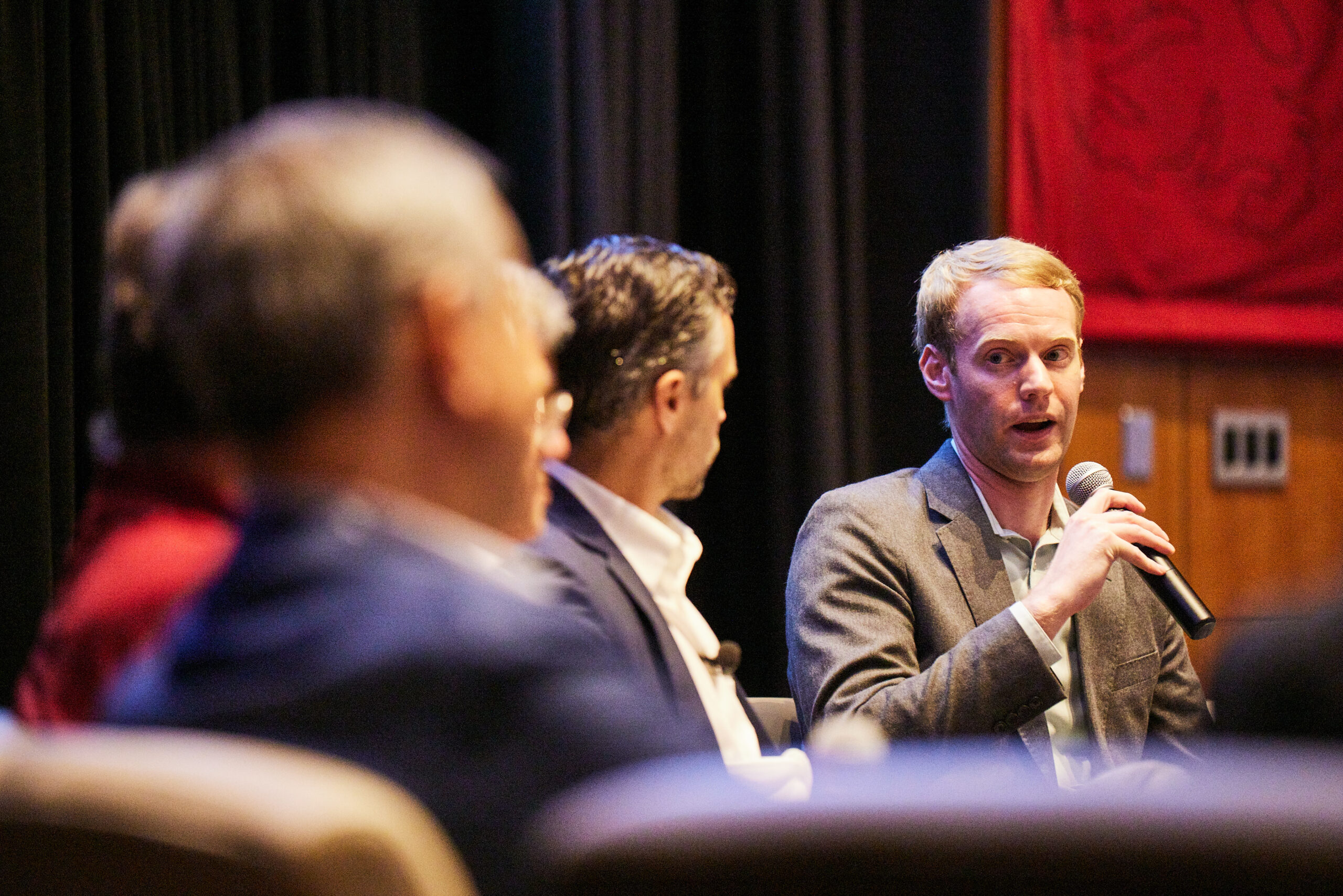 Andrew Kruse speaks into a microphone while seated next to four other pictured panelists.