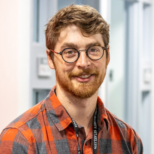 Man smiles in office for a headshot. He wears glasses and has a beard.