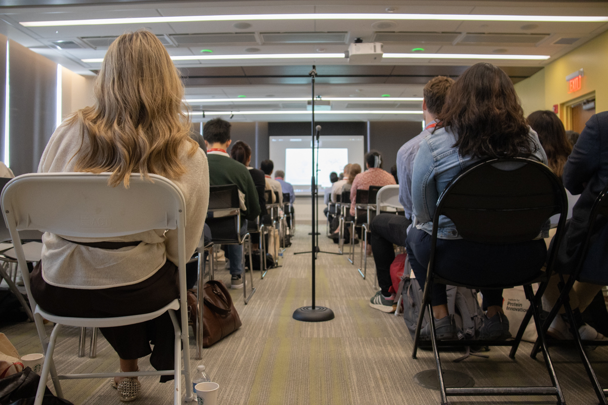 The image looks down the aisle toward a screen, with a microphone in the middle of the aisle. Either side of the aisle is lined with people sitting in folding chairs watching the presentation.