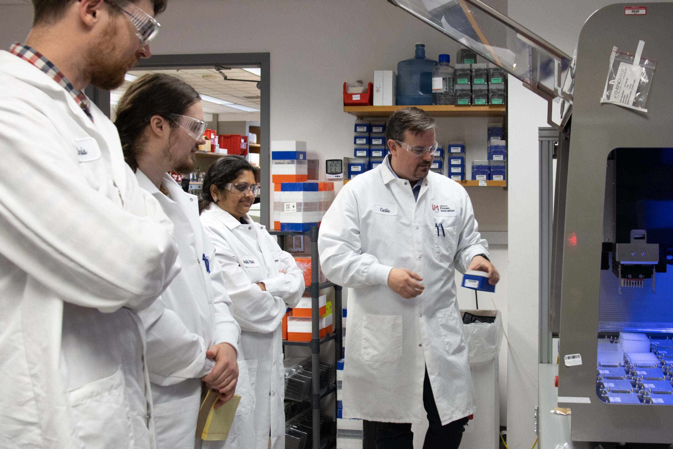 A man in a lab coat and safety glasses adds a plate of antibody samples to a robot in a lab. A group of three scientists stand in a line, looking at the machine.