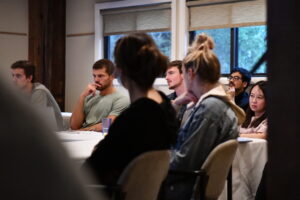 Seven scientists sit around the corner of a table, listen intently to a talk. Two are silohetted in the foreground.