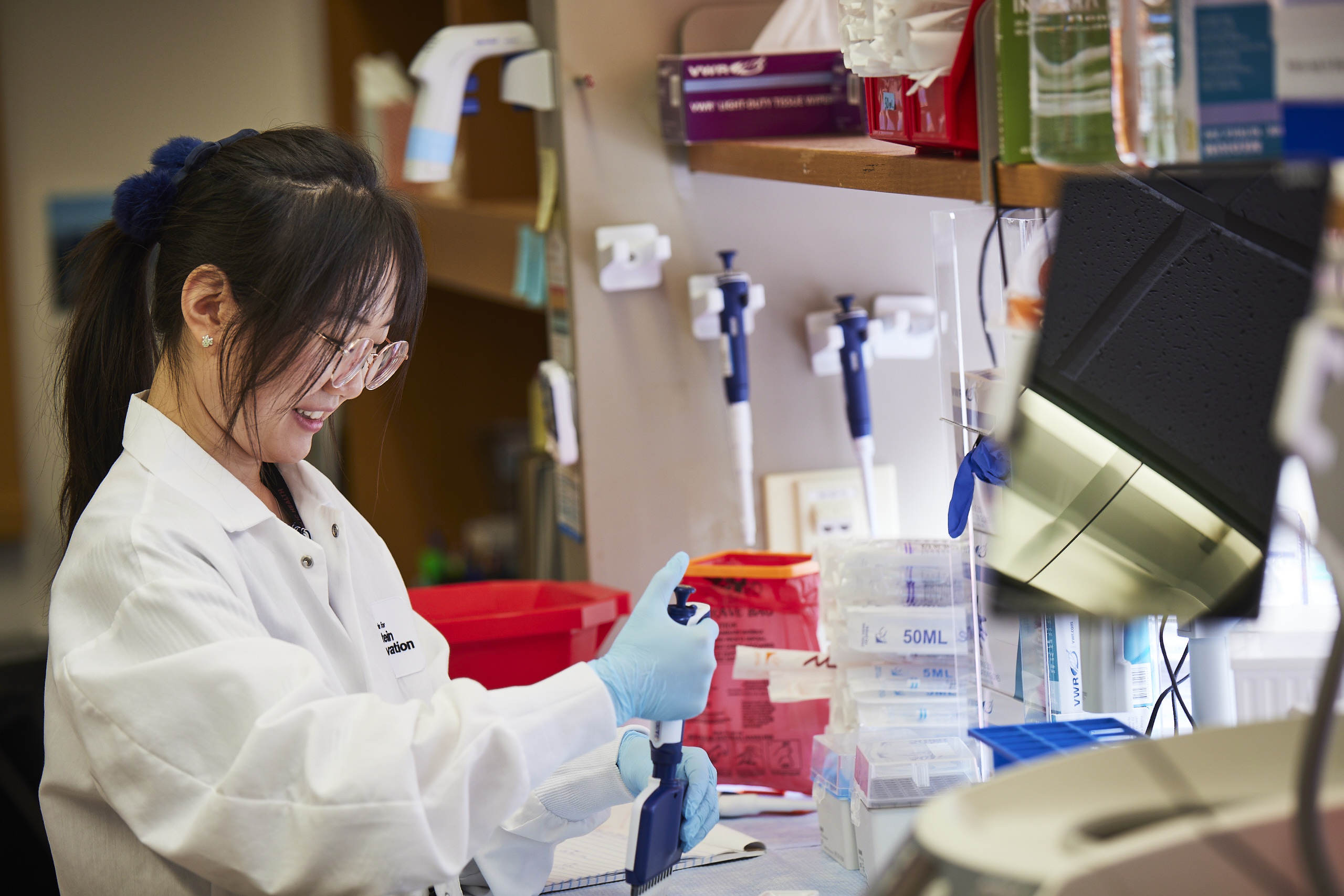 A woman, wearing eye glasses, a lab coat and gloves, uses a multi-channel pipette at a lab bench.