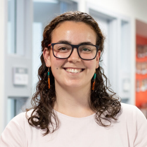 Rebecca smiles in the IPI office. She wears glasses and earrings, and has curly hair draped over her shoulders.