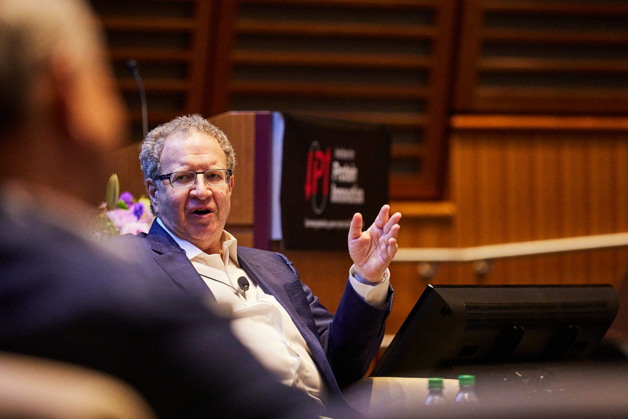 Ken Fasman sits on stage in front of an IPI sign, motioning his hands while mid-sentence.