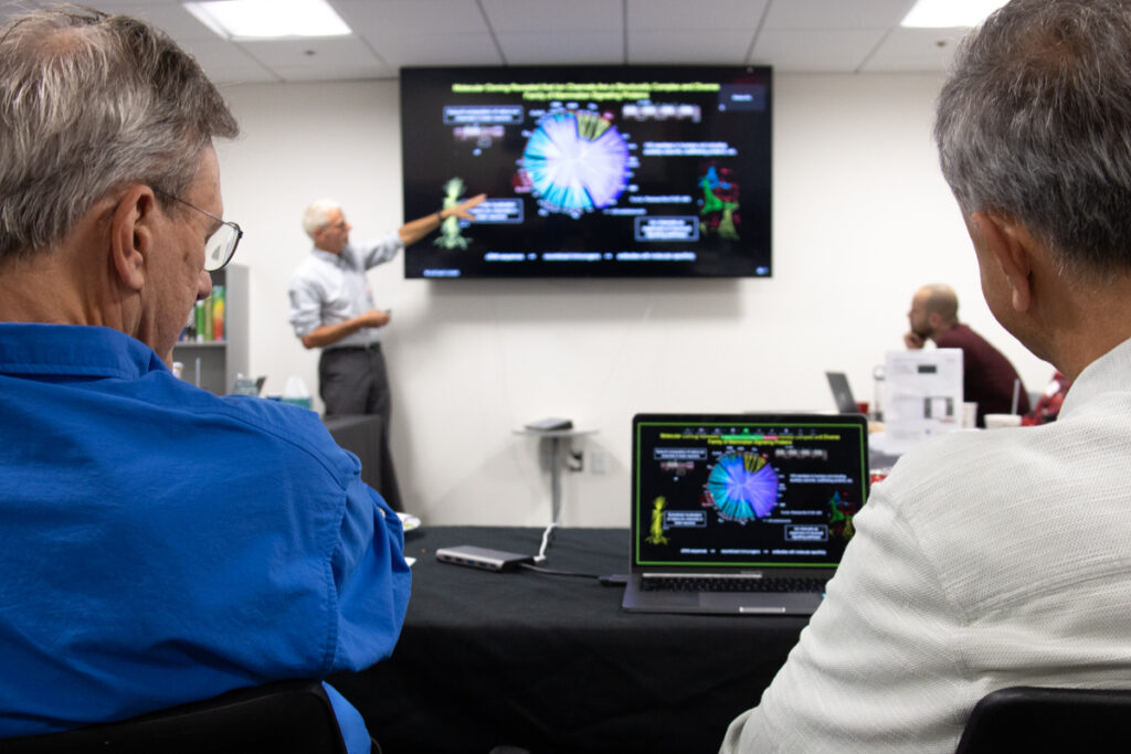 Two scientists sit at a table, watching a presentation. Viewable between them, a scientist at the front of the room points to a screen as they give a presentation about antibodies.