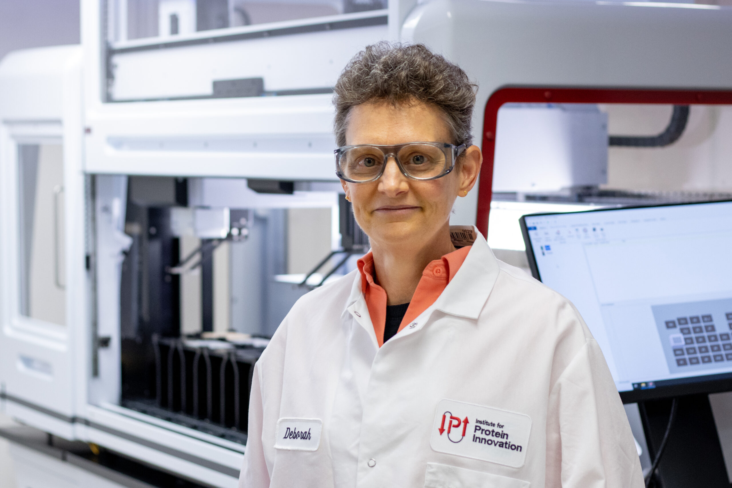 Deb Moshinsky stands in front of a lab machine wearing an IPI lab coat and safety goggles.