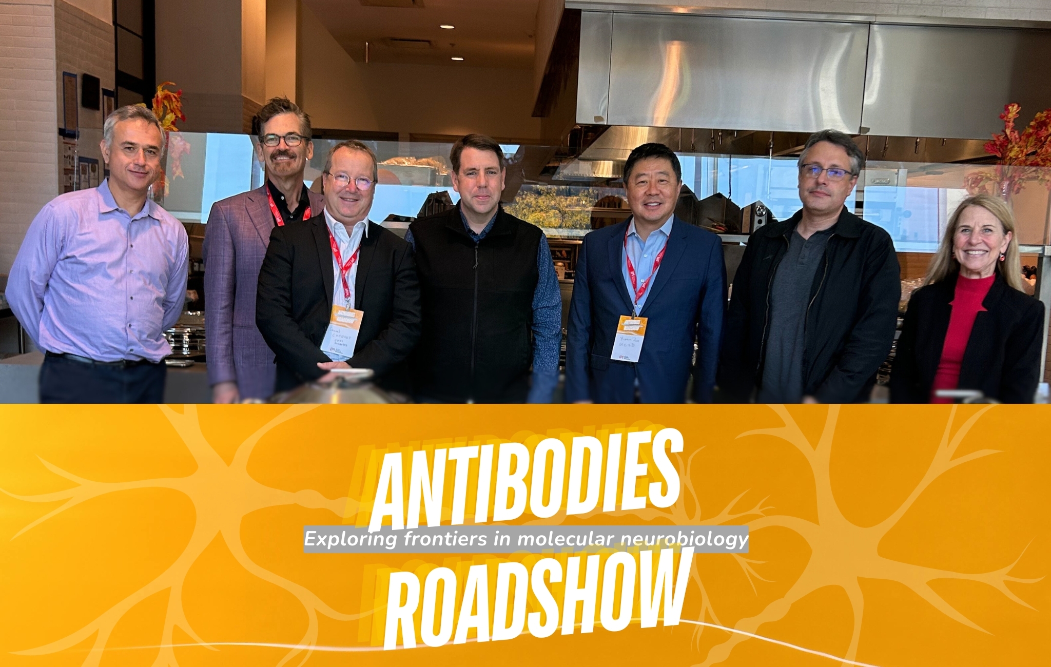 A group of scientists wearing lanyards pose for a photo after lunch at a scientific conference. A graphic at the front of the image reads, "Antibodies Roadshow: Exploring frontiers in molecular biology."